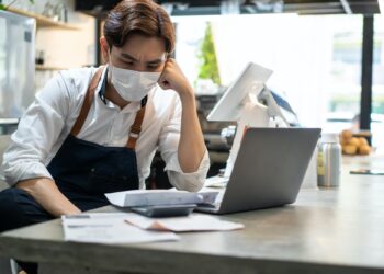 Depressed Asian cafe owner wear protective mask feel worried about financial problem in coffee shop